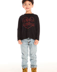 Full body view of boy wearing Chaser’s Heartbreaker black long-sleeve shirt with red text, broken heart graphic, light blue jeans, and brown boots on a white background