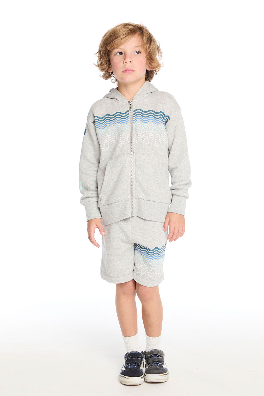 Full body view of boy wearing the Chaser Kids Peace Out Shorts in Heather Grey styled with the matching Peace Out Zip-Up Hoodie featuring blue wave graphics across the chest and shorts, paired with white socks and navy sneakers on a white background