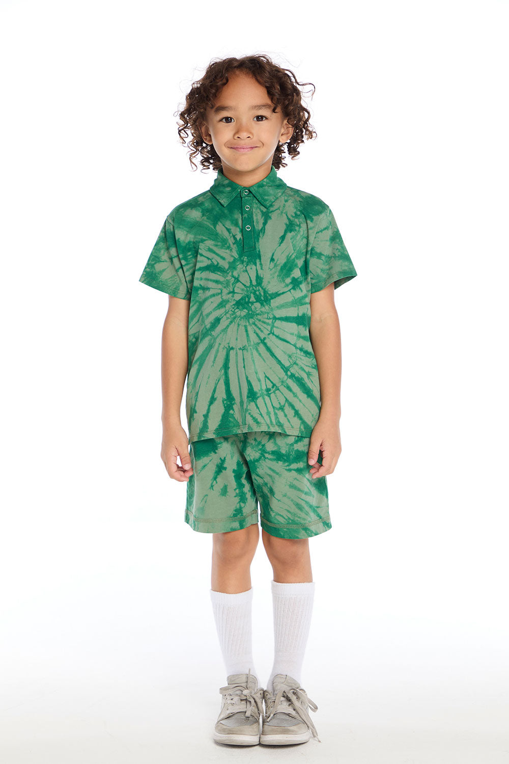 Full body view of boy wearing the Chaser Kids Wild Green Tie Dye Polo Shirt and matching Wild Green Tie Dye Cozy Beach Shorts styled with white socks and silver sneakers on a white background