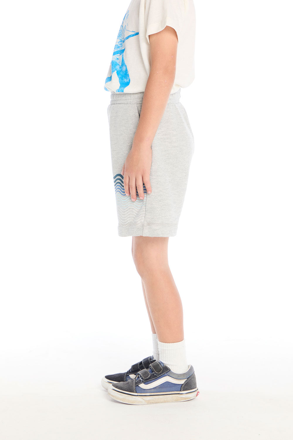 Side view of boy wearing the Chaser Kids Peace Out Shorts in Heather Grey showing side pockets and blue wave design, styled with a graphic tee and navy sneakers on a white background