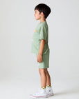 Side view of boy wearing Chaser Kids Lets go Golfing shorts and matching green t-shirt with white sneakers on a white background