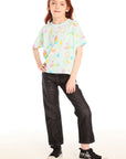 Full body view of girl wearing Chaser’s light green Unicorn Hearts Tee with text and colorful graphics, black jeans, and shiny sneakers against a white background