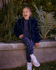 Chaser Kids Avalon Girls Puff-Sleeve Jacket in navy, full-body view of a girl seated on a low wall outdoors beside greenery