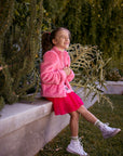 Chaser Kids Camelia Girls Puff-Sleeve Jacket in pink, full-body view of a girl seated on a low wall outdoors beside greenery