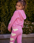 Chaser Kids Holiday Hug Girls Hoodie in pink, rear three-quarter view with hood down outdoors beside a low wall and greenery