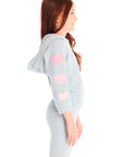Side view of girl wearing Chaser’s light blue Cropped Shirred Snowflake Pullover Hoodie with pink hearts on the sleeves and matching Slim Snowflake Legging pants on a white background