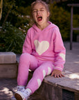 Chaser Kids Holiday Hug Girls Pants in pink with white hearts, worn by a girl seated on a low wall outdoors beside greenery