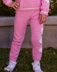 Chaser Kids Holiday Hug Girls Pants in pink with white hearts down one leg, front view outdoors beside a low wall and greenery