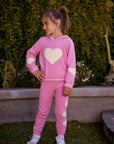 Chaser Kids Holiday Hug Girls Hoodie and Holiday Hug Girls Pants in pink with white hearts, full-body front view outdoors beside a low wall and greenery