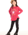 Full body view of girl wearing Chaser’s red Hot Chocolate Weather Long Sleeve with a graphic design, black pants, and a brown hat on a white background