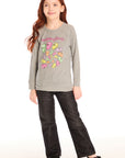 Full body view of girl wearing Chaser’s gray The Rolling Stones Spiral Tongues Long Sleeve with colorful logo designs and text, black jeans, and sparkly shoes on a white background