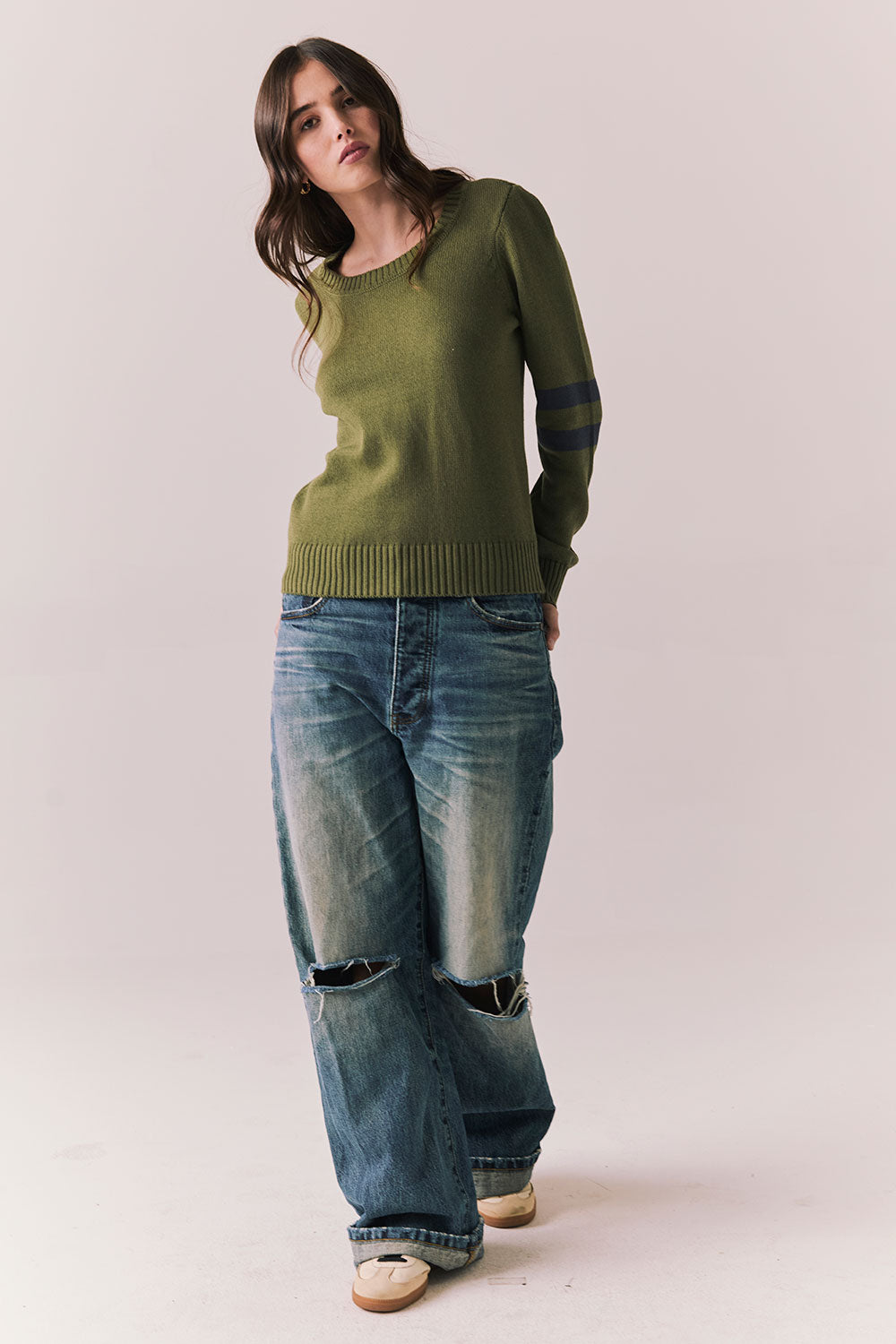Full front view of model wearing the Varsity Star Sweater in bronze green with slouchy fit and twin arm stripes and blue jeans on a plain background
