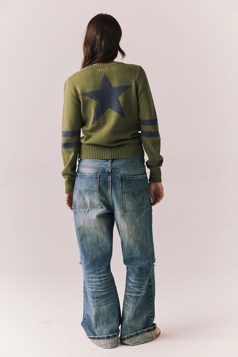 Back full body view of model wearing Varsity Star Sweater in bronze green with large star graphic on back and twin arm stripes and blue jeans on a plain background