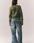 Back full body view of model wearing Varsity Star Sweater in bronze green with large star graphic on back and twin arm stripes and blue jeans on a plain background