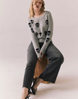 Woman wearing the Tossed Skulls Long Sleeve sweater in Heather Grey with allover black skull motif and fitted silhouette sitting on a wooden block against a plain background.