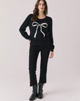 Full body view of woman wearing Chaser’s true black Bow Long Sleeve with white bow design, black jeans, and boots on a plain background.