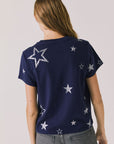 Back view of woman wearing Chaser's Allover Distressed Stars Tee with star patterns and dark denim pants on a plain background