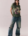 Woman wearing the Woodstock Festival Flower Tee in bronze green with cream and pink floral graphic and vintage font with blue denim against a plain background