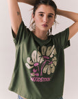 Front view of model wearing Woodstock Festival Flower Tee in bronze green with cream and pink floral graphic and vintage font against a plain background