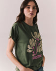 Side view of model wearing Woodstock Festival Flower Tee in bronze green with cream and pink floral graphic and vintage font against a plain background