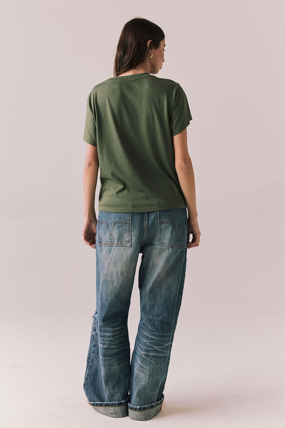 Back view of woman wearing the Woodstock Festival Flower Tee in bronze green with cream and pink floral graphic and vintage font with blue denim against a plain background