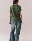 Back view of woman wearing the Woodstock Festival Flower Tee in bronze green with cream and pink floral graphic and vintage font with blue denim against a plain background