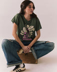 Woman wearing the Woodstock Festival Flower Tee in bronze green with cream and pink floral graphic and vintage font with blue denim sitting on a wooden block against a plain background