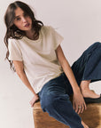 Woman sitting on a wooden box wearing the Rambler Starry White Tee with ringer neck and cap sleeves and blue jeans against a plain background