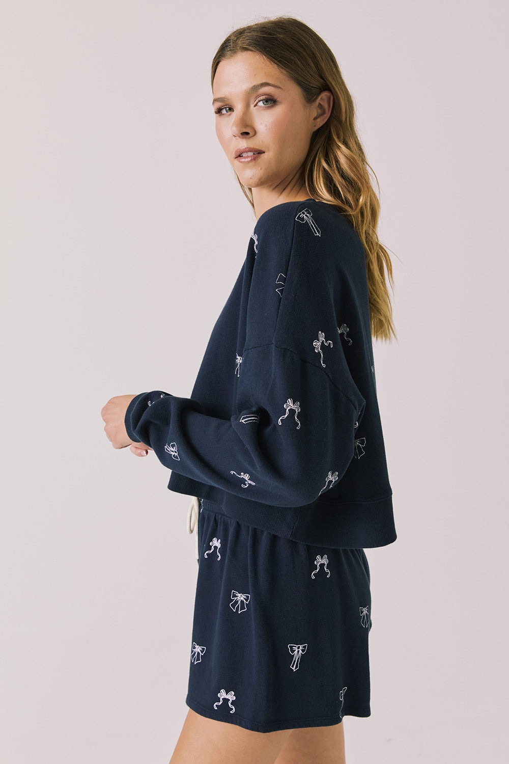 Side view of woman wearing Chaser’s navy Tossed Bows Long Sleeve with bow pattern on a white background