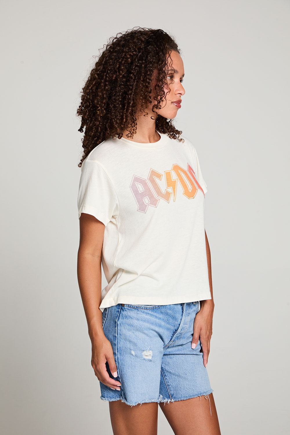 Side view of model in AC/DC 1979 Tour Gradient Tee styled with denim shorts for a retro festival look against a plain background