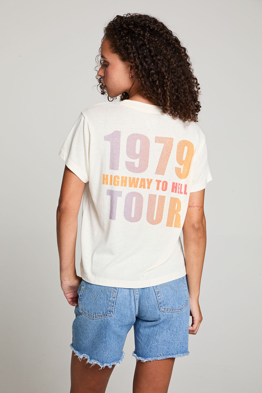 Back view of model in AC/DC 1979 Tour Gradient Tee styled with denim shorts for a retro festival look against a plain background
