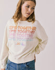 Front view of woman wearing Chaser’s The Beatles Come Together Starry White Long Sleeve with ‘COME TOGETHER’ text in rainbow colors on a plain background