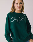 Front view of woman wearing Chaser’s green Cin Cin! Long Sleeve with “Cin Cin” text and star graphics on a plain background