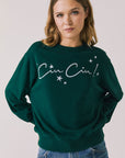 Front view of woman wearing Chaser’s green Cin Cin! Long Sleeve with “Cin Cin” text and star graphics on a plain background