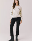Full body view of woman wearing Chaser’s Starry White French Scripts Long Sleeve with French phrases like “Merci” and “Bonjour” in black text, black pants, and black boots on a white background