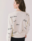 Back view of woman wearing Chaser’s Starry White French Scripts Long Sleeve with French phrases like “Merci” and “Bonjour” in black text on a white background