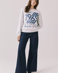 Full body view of woman wearing Chaser’s light grey USA Sports Club Long Sleeve with a blue USA 1969 graphic and dark jeans on a white background