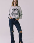 Full body front view of a woman wearing Chaser's gray Health Is Wealth Long Sleeve with a graphic design and ripped denim jeans on a plain background
