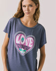 Front view of woman wearing Chaser’s slate blue Woodstock Love Revolution Tee with pink heart design and ‘LOVE’ text on a plain background (licensed music collaboration)