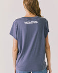 Back view of woman wearing Chaser’s slate blue Woodstock Love Revolution Tee with Woodstock in white text on a plain background (licensed music collaboration)