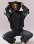 Full body view of model wearing the Halftime fleece pullover hoodie in True Black with ‘HALFTIME’ chest graphic, crouching on the floor against a plain background.