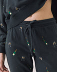 Front view of woman wearing Chaser’s black Champagne Party Joggers with allover champagne bottle and glass graphics on a light grey background