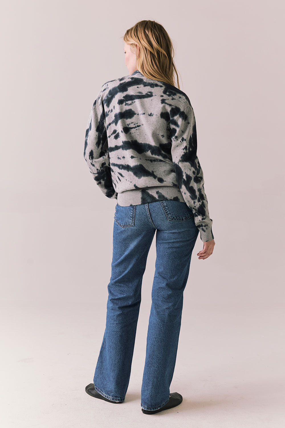 Back full body view of model in black tie dye hoodie and blue jeans on a plain background