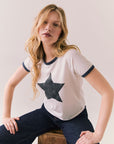 Woman wearing a white t-shirt with a star design, sitting on a wooden stool against a plain background.