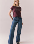 Full front view of model in J’adore Vinyl Tee in fig purple with cream ringer trim and vintage-style text and blue jeans on a plain background