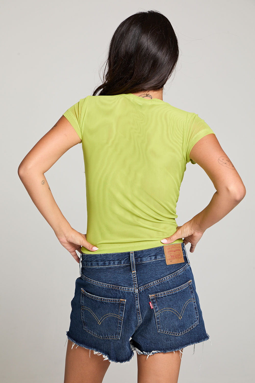 Back view of model wearing the 90’s Chartreuse Baby Tee in bright green with scoop neck and fitted cropped cut and cut off denim against a plain background