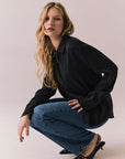 Full side view of a model wearing the Helenaa button-down shirt in True Black with collared neckline and oversized silhouette, styled with relaxed jeans and crouched against a plain backdrop.