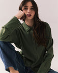 Front view of a model wearing the Helenaa button-down shirt in bronze green with collared neckline and relaxed silhouette, styled with blue jeans, sitting on the floor against a plain background.