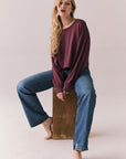 Full view of model wearing the Hartt long sleeve tee in fig (deep purple) with clean silhouette and blue jeans, seated on a wooden block against a plain background.