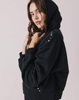 Side view of woman wearing Chaser’s Kiki True Black Zip Up Hoodie with sequin embellishments, hood up, and matching Daze True Black Joggers on a white background